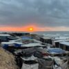 Improvised camp for displaced people on the coast of the Gaza Strip at sunset, with tents and shelters made of tarpaulins and assorted materials, clustered closely under a cloudy sky. In the background, the sea and the sun are partially hidden behind grey clouds. Gaza